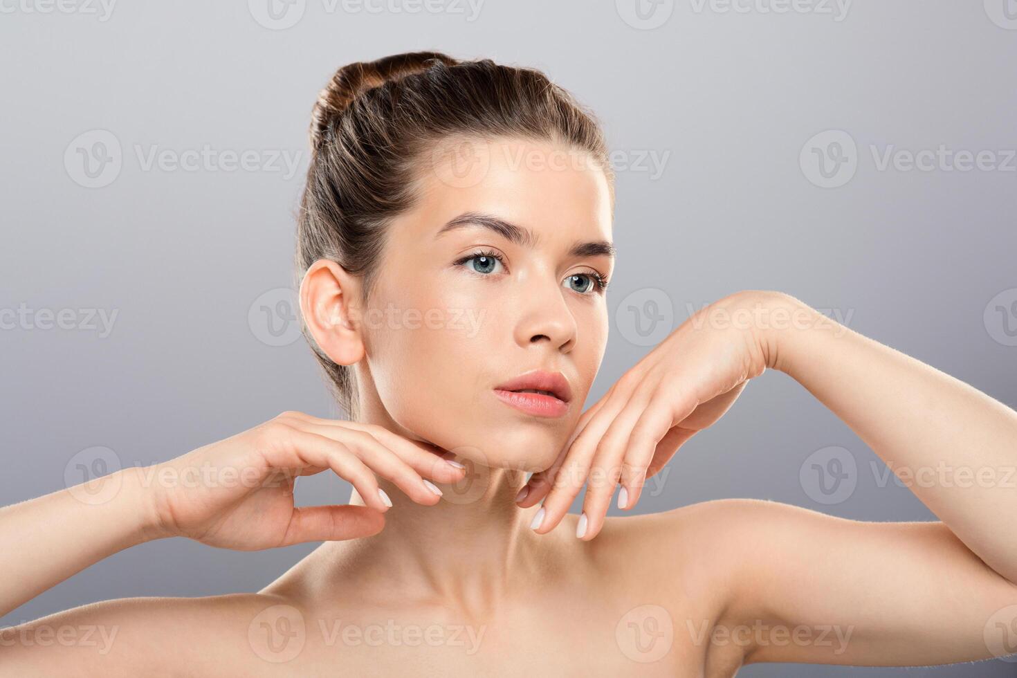 Beautiful sensual young woman posing over grey studio background, touching her pretty face, female beauty concept. Attractive nude lady on gray, touching her chin with both hands, copy space photo