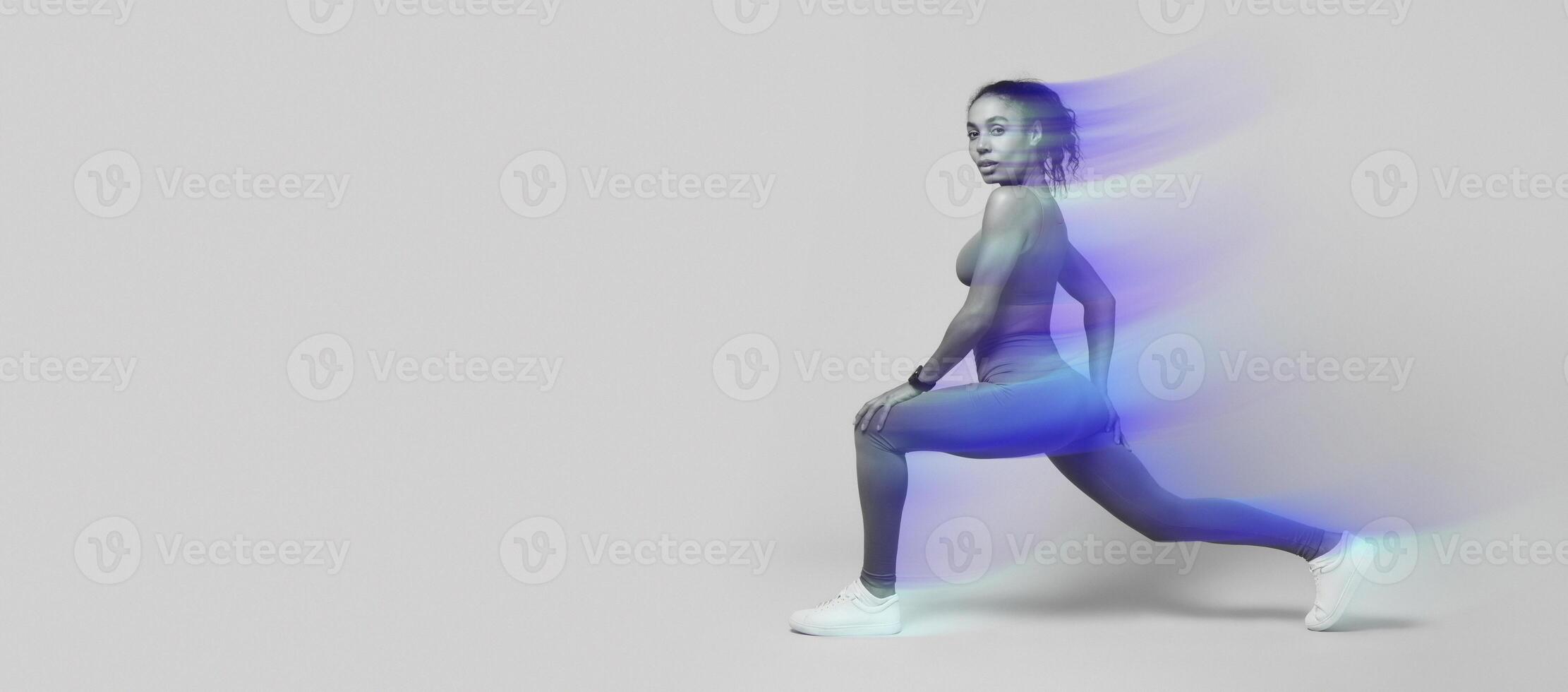 Sporty African American woman lunging forward exercising during workout over pink studio background, looking at camera with confidence. Gym training for muscles flexibility. Side view shot photo