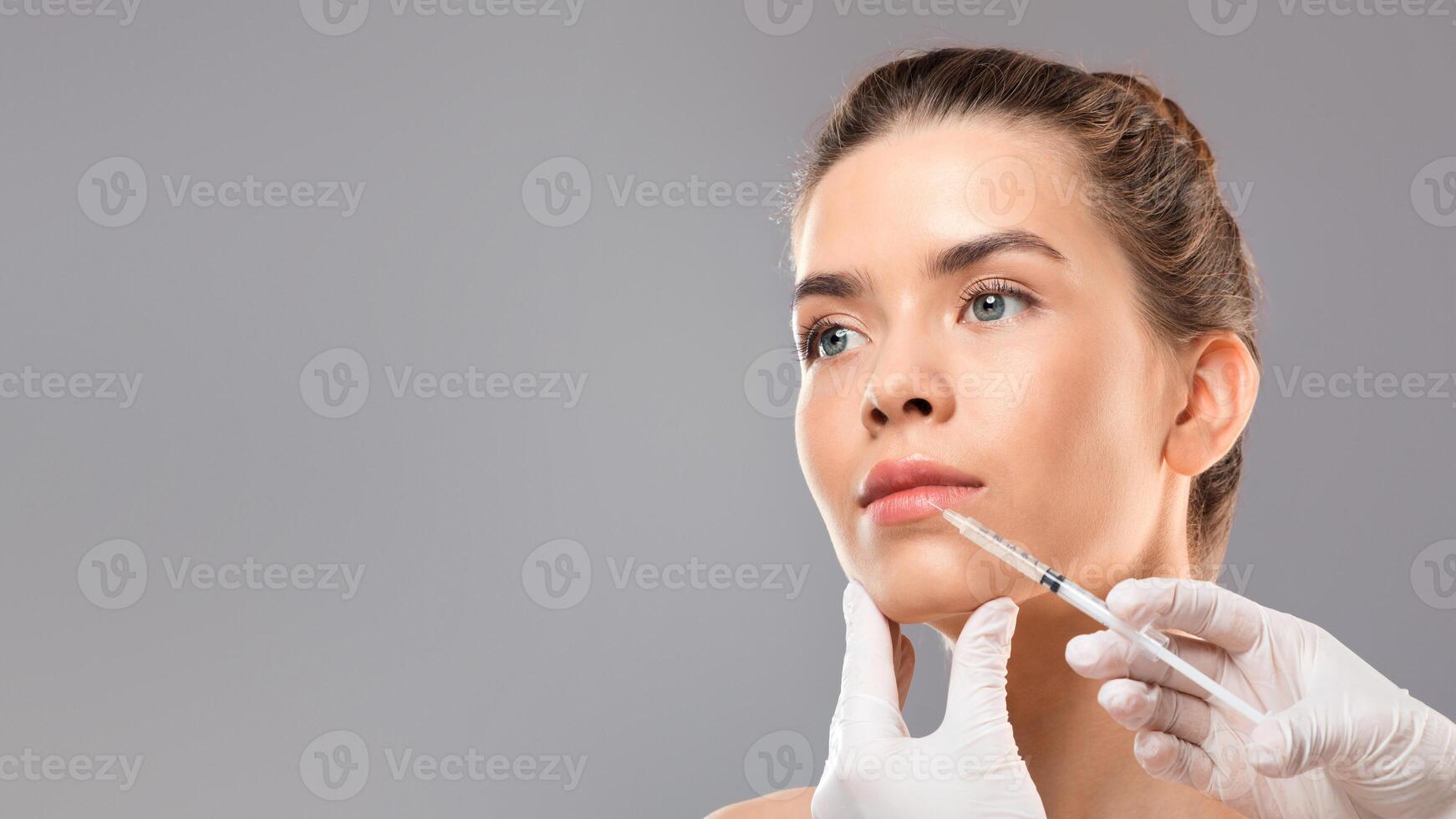 Aesthetic modern cosmetology concept. Lip injection for young woman over grey studio background, closeup. Unrecognizable cosmetitian injecting lip filler for beautiful lady looking at empty space photo