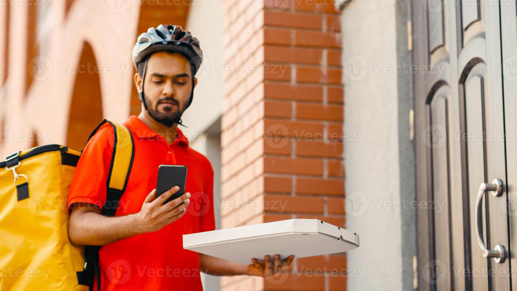 Pizza delivery with man on bike. Serious delivery man with beard in helmet, with yellow backpack holding box and looking at smartphone, near front door, free space photo