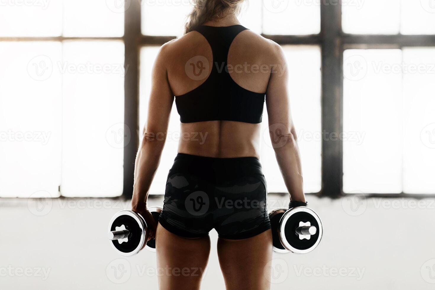 Strength training at home or gym during lockdown, self-isolation and pandemic. Muscular adult female athlete in sportswear does exercise with dumbbells for arms in loft interior with window, back photo