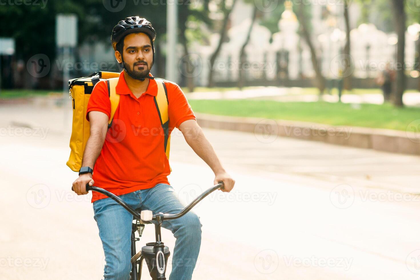 Portrait of male courier, riding on bicycle with delivery backpack. Young guy in protective helmet on road in city, panorama, sun flare, free space photo