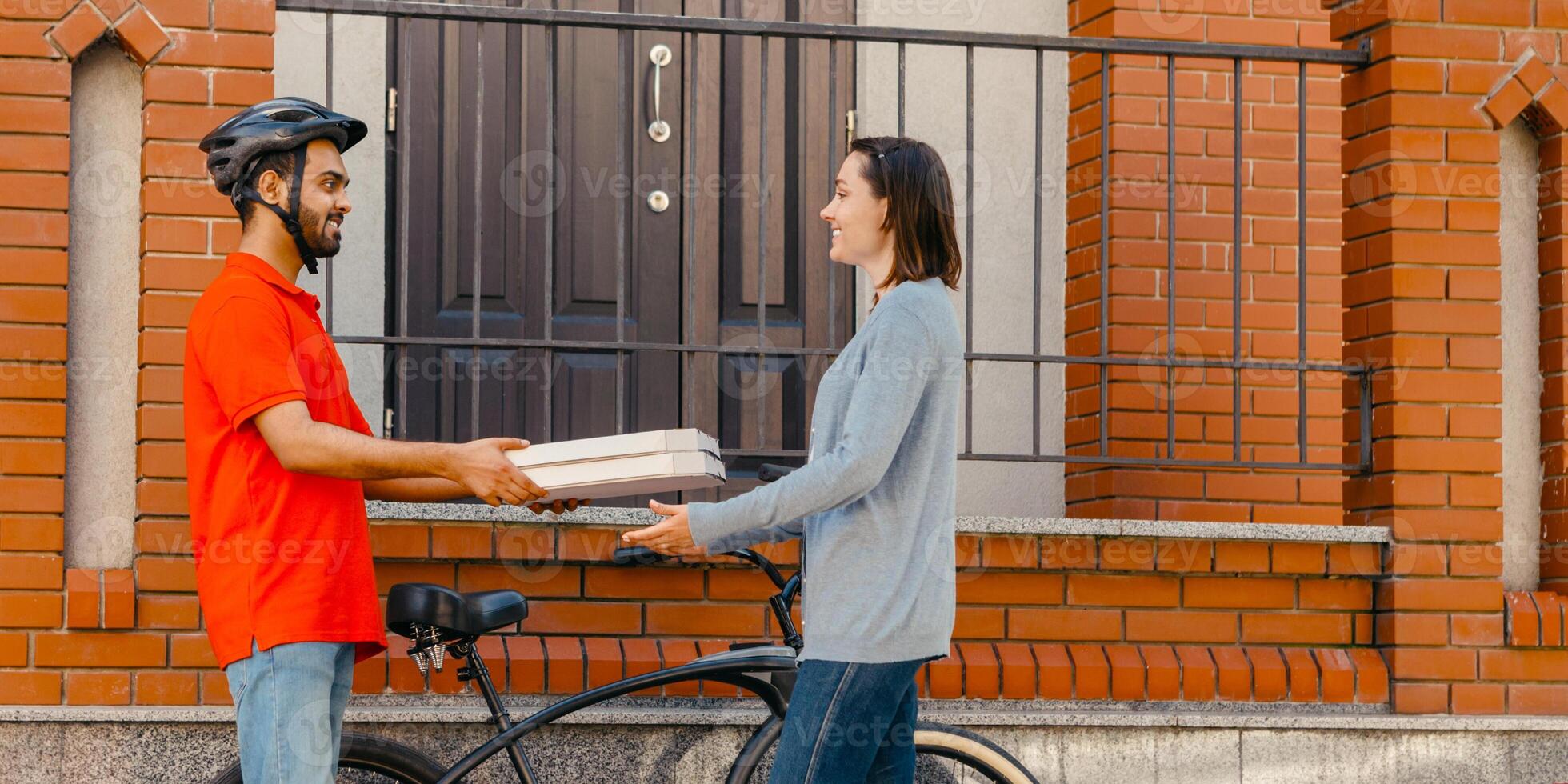Ecological city, bike and delivering service. Smiling courier in helmet gives pizza to client, near entrance, at street, side view, free space photo