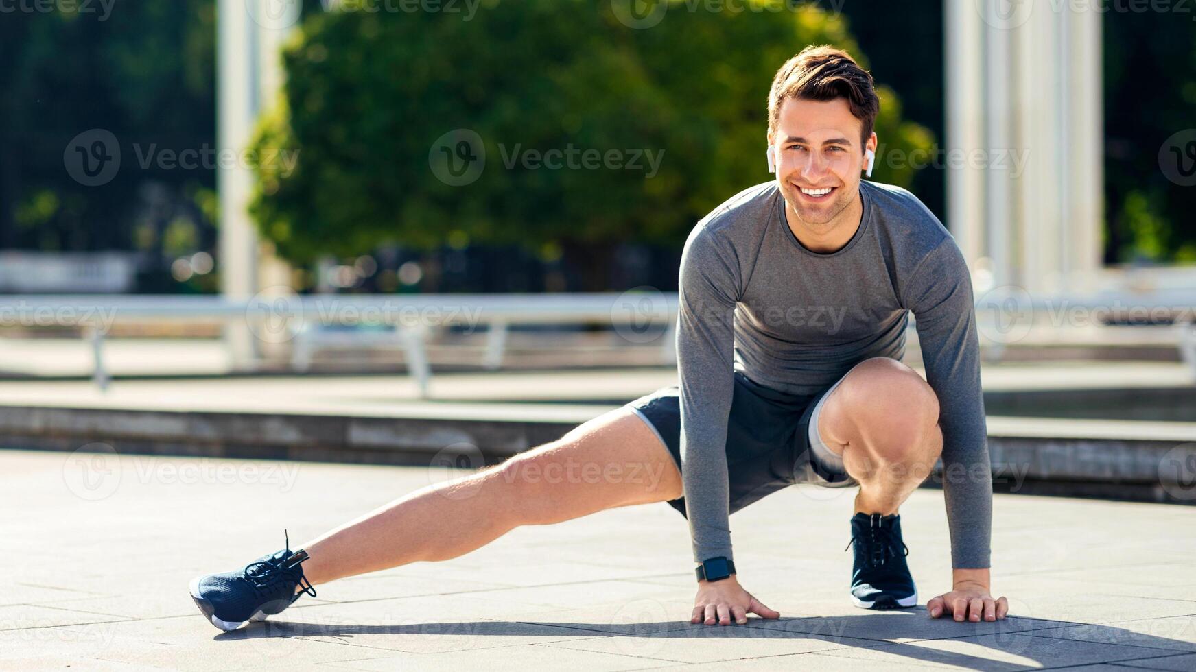 Warm up before run in park. Young smiling man in sports uniform doing leg exercises, outdoor, free space photo
