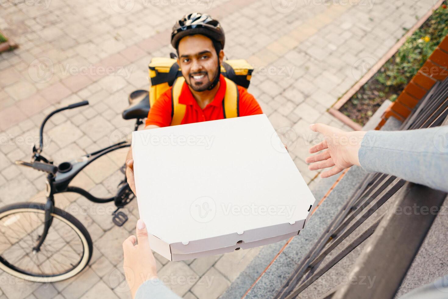 Online order and pizza at home. Smiling courier with beard in helmet gives boxes to client, near house on street, top view, pov photo