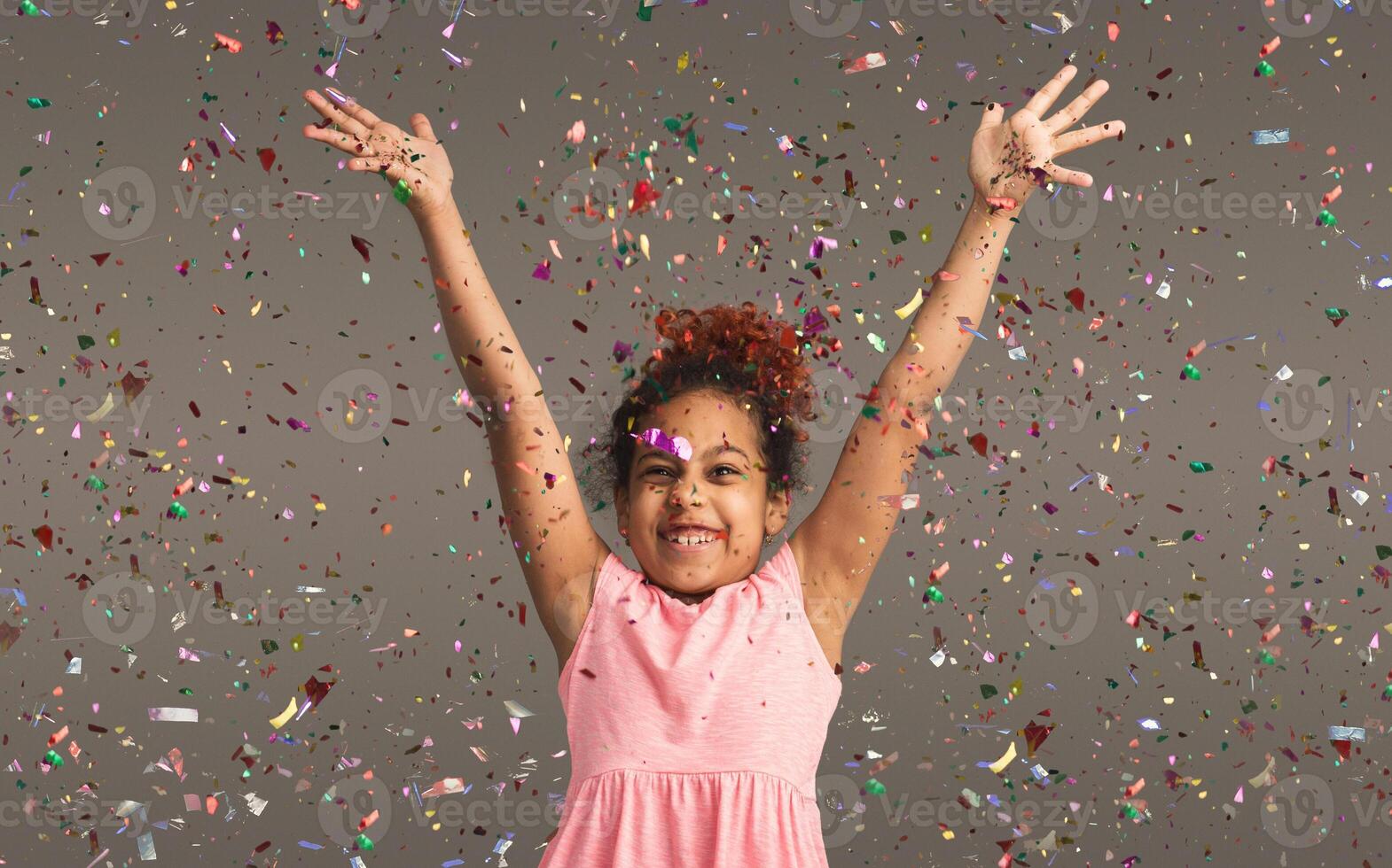 Happy african-american little girl at gray studio background, copy space. Portrait of beautiful child having fun with shiny confetti photo