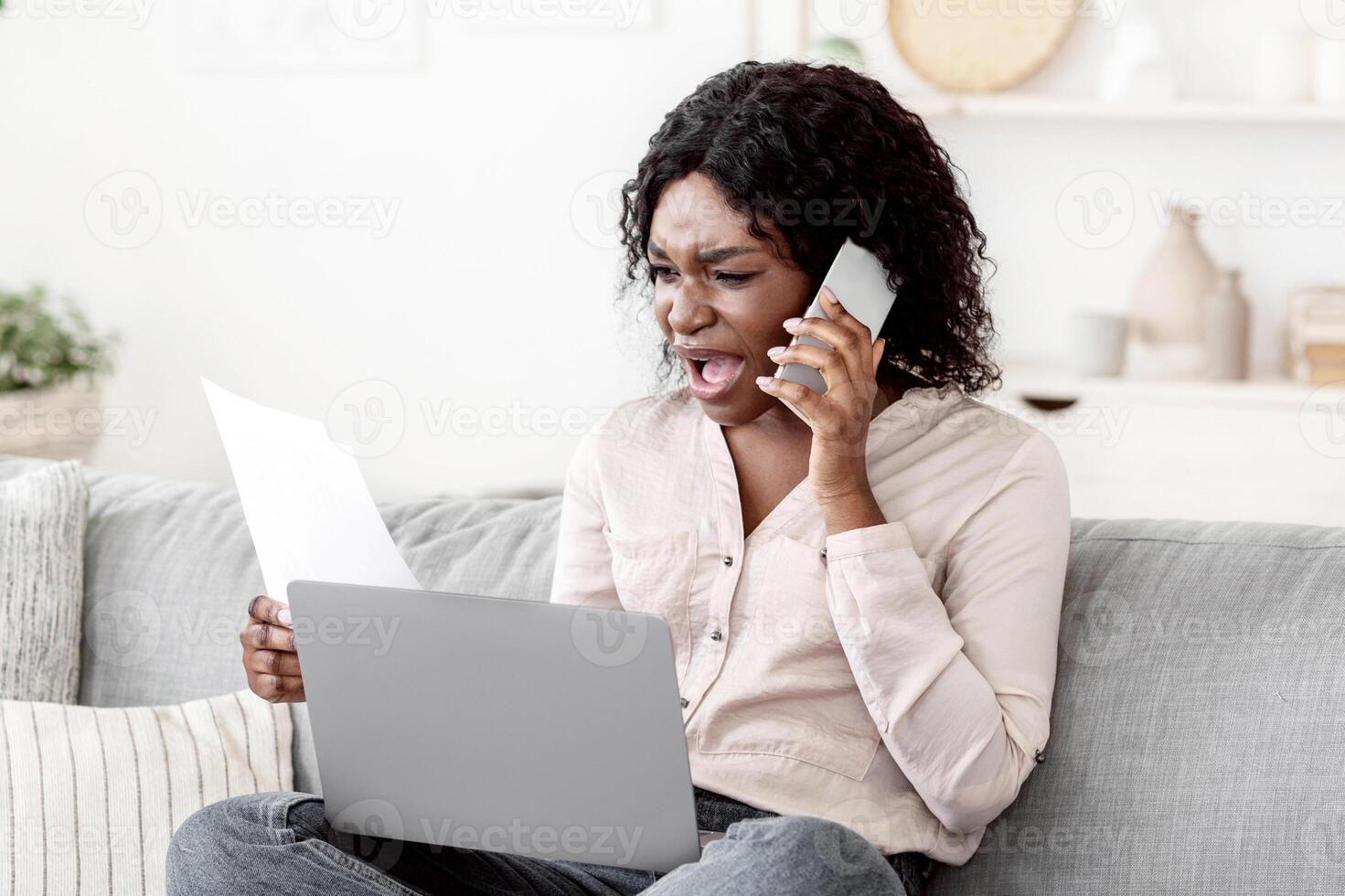 Remote Work Stress. Angry black woman having problems working with laptop and papers at home, checking documents and emotionally talking on cellphone, sitting on couch in living room, free space photo
