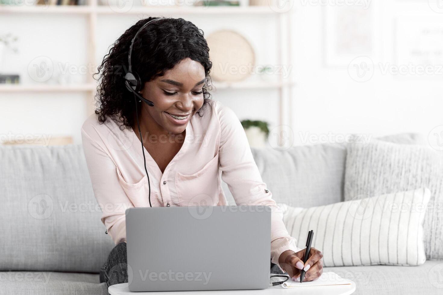 Distance Learning. Positive Black Woman Studying With Laptop And Headset At Home, Watching Webinar And Taking Notes To Notepad photo