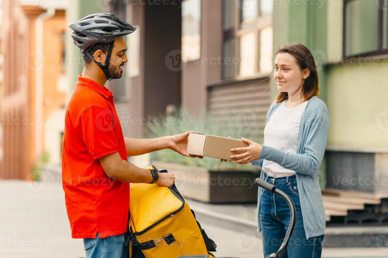 Satisfied customer receives online order. Girl takes cardboard parcel from courier with yellow backpack and bicycle on street, side view, free space photo