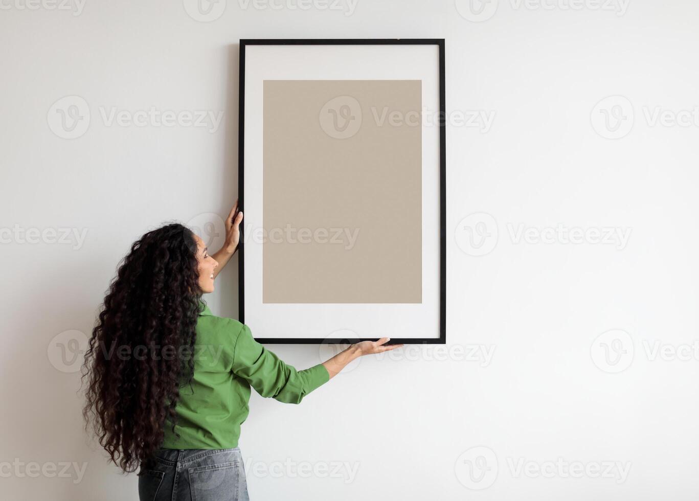 Happy Woman Hanging Empty Poster In Frame On White Wall Standing Indoors. Modern Art, Domestic Interior And Home Decor Pictures Concept. Mockup For Advertisement photo