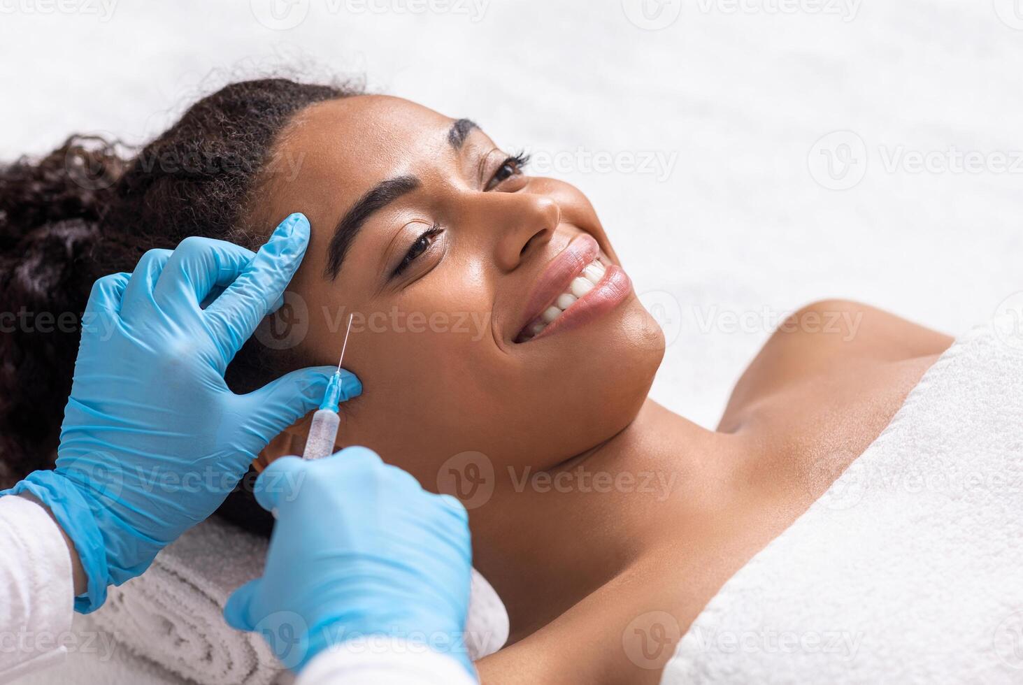 Smiling black woman attending beautician, having anti-aging injection for eye zone, closeup photo