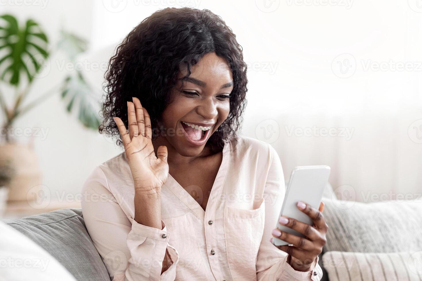 Joyful African Woman Having Call With Friends Via Smartphone At Home, Waving Hand At Camera, Greeting Somebody, Empty Space photo