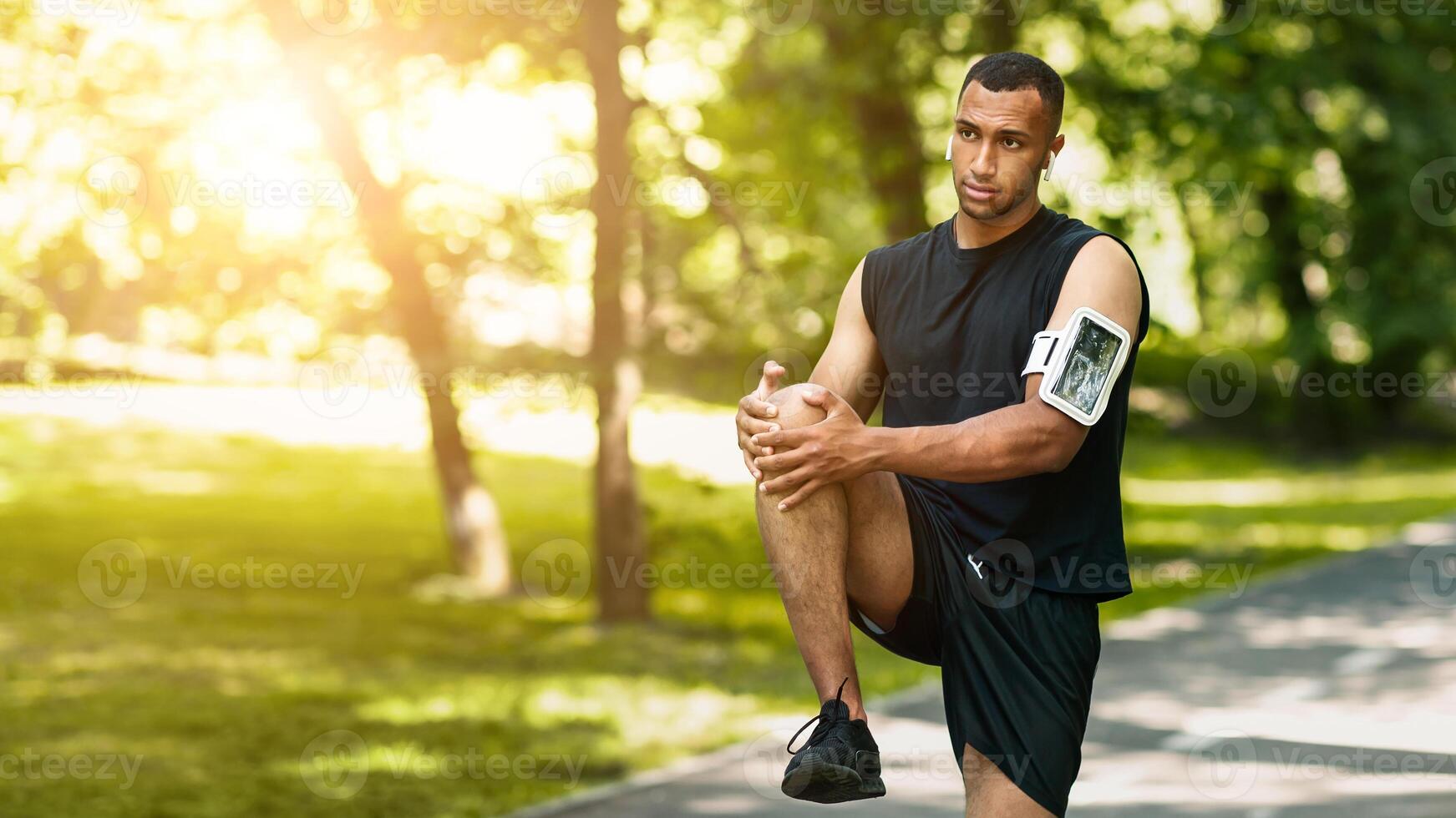 Handsome black athlete stretching his leg after jogging at summer park, panorama photo