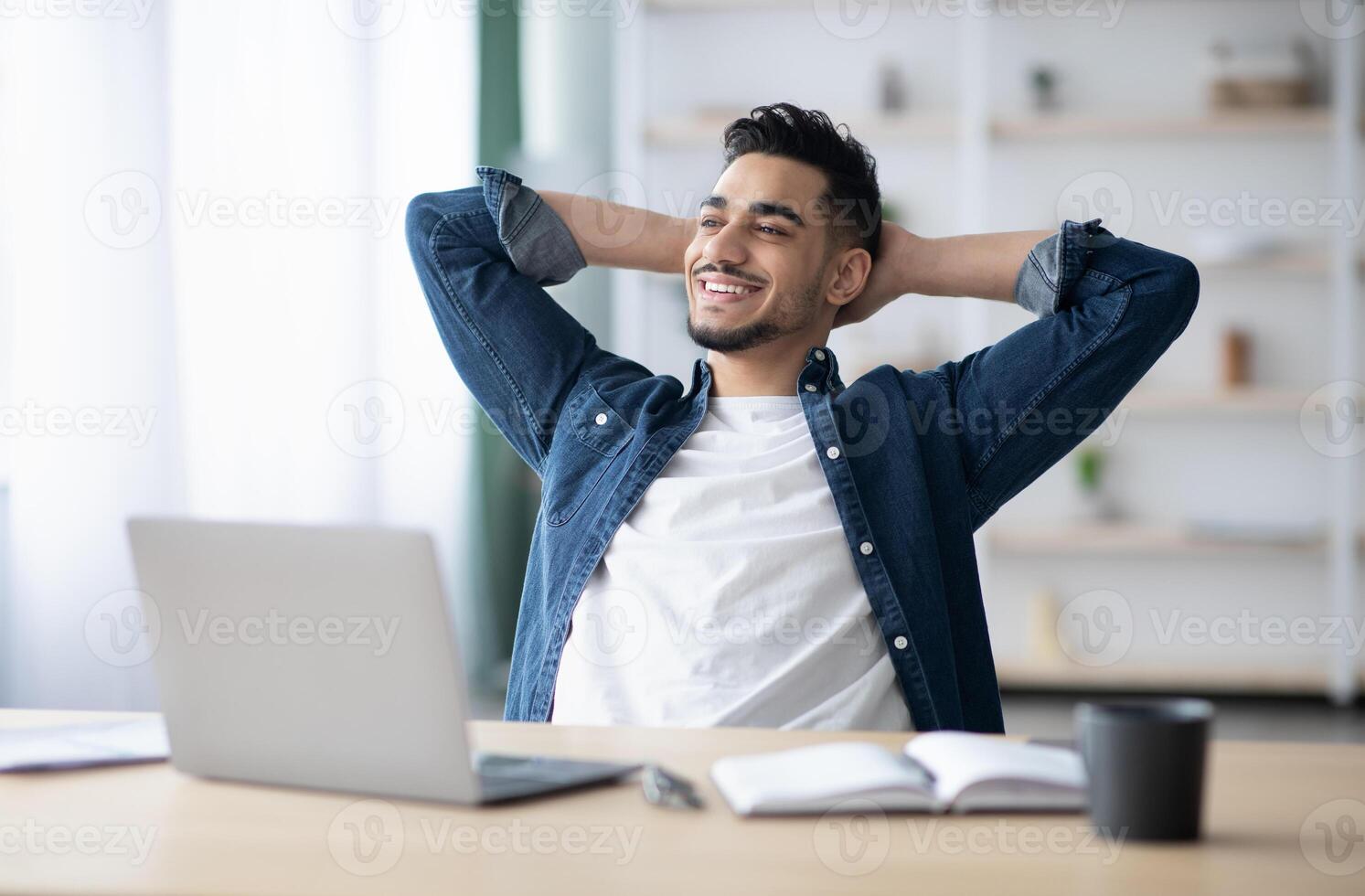 Happy young arab man enjoying his job, sitting at workdesk, leaning back, looking at copy space and smiling, office interior. Job opportunities for millennials, career concept photo