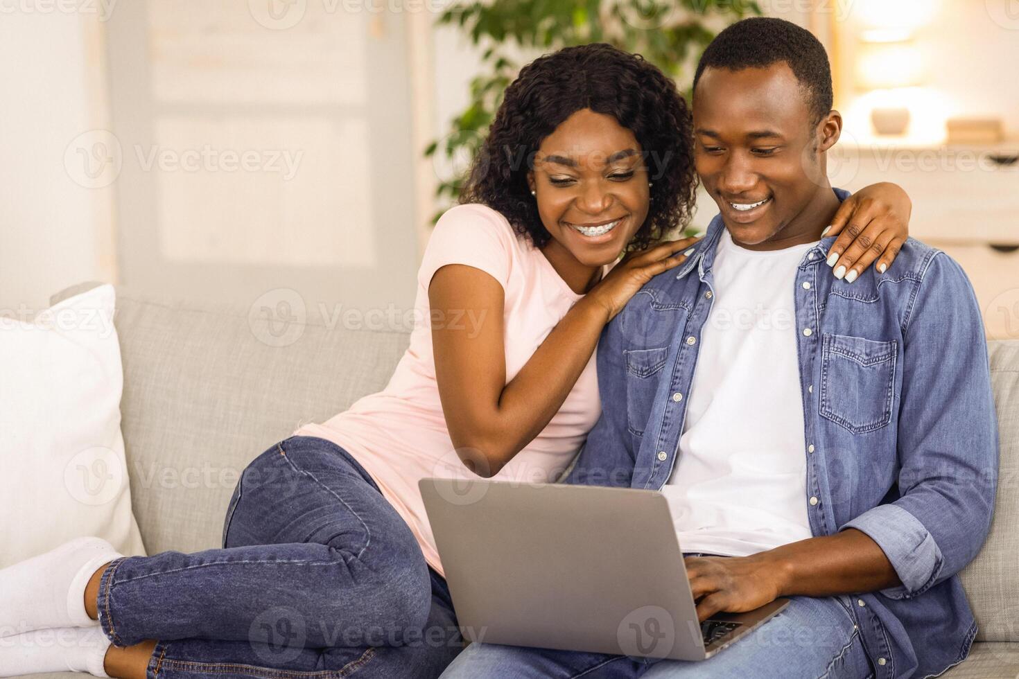 Cheerful black couple using laptop while resting on couch at living room at home, watching funny content together, copy space. African american loving family husband and wife spending time together photo