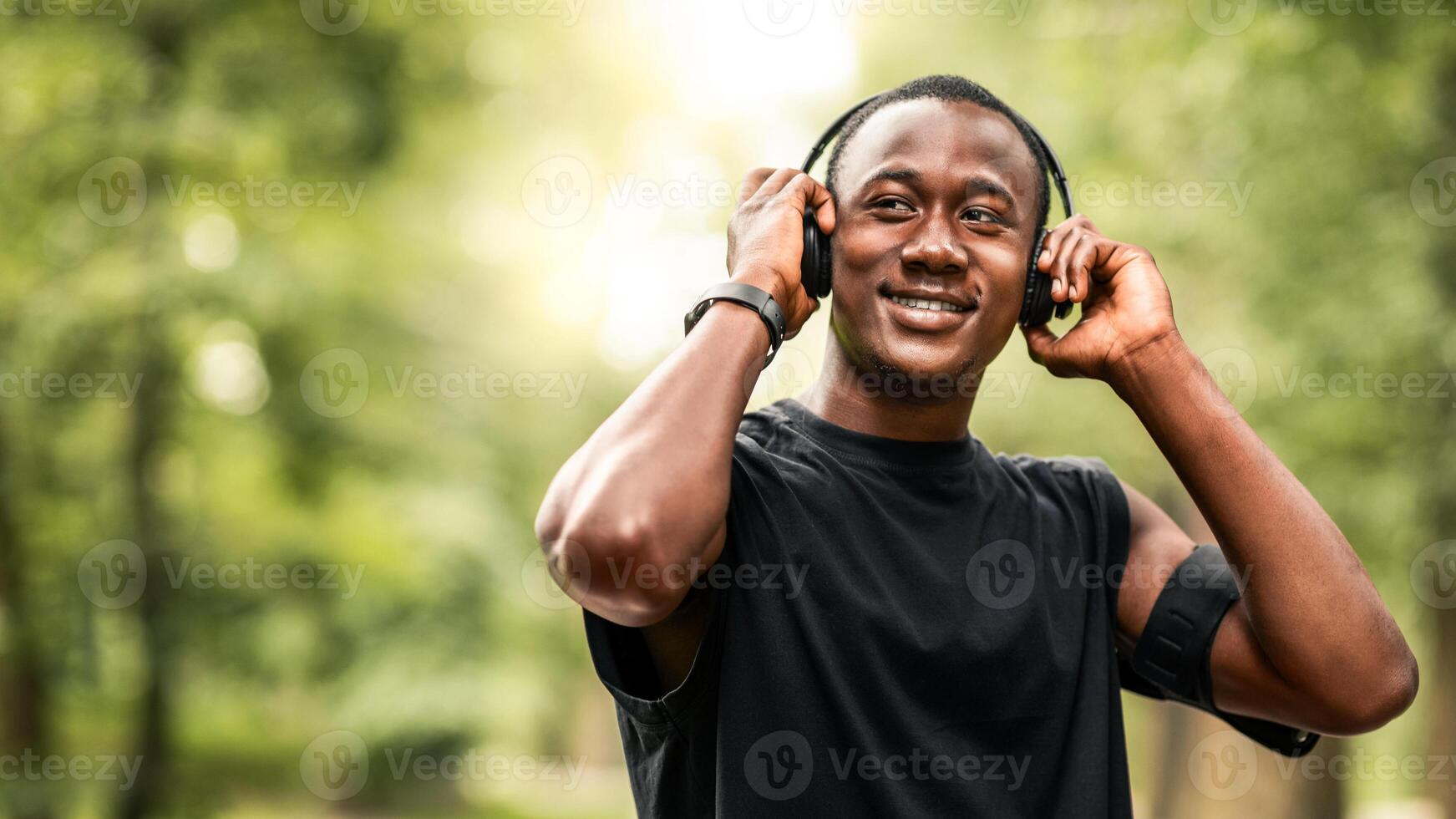 Music for exercising. Joyful black jogger putting wireless headset on his head, training at park, looking at free space photo