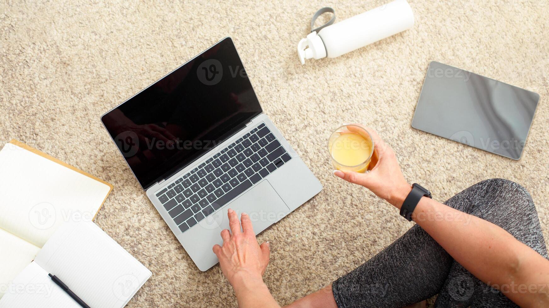 Healthy lifestyle, blog and sport. Woman with smart watch holds glass of orange juice and works on laptop, sitting on floor, with notepads, tablet and bottle, cropped, free space photo