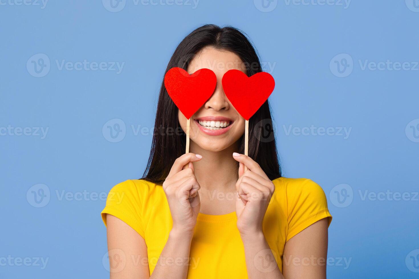 All You Need Is Love. Romantic asian woman posing with red hearts over eyes and smiling, having fun, celebrating valentine's day alone, standing over purple studio background, closeup shot, free space photo