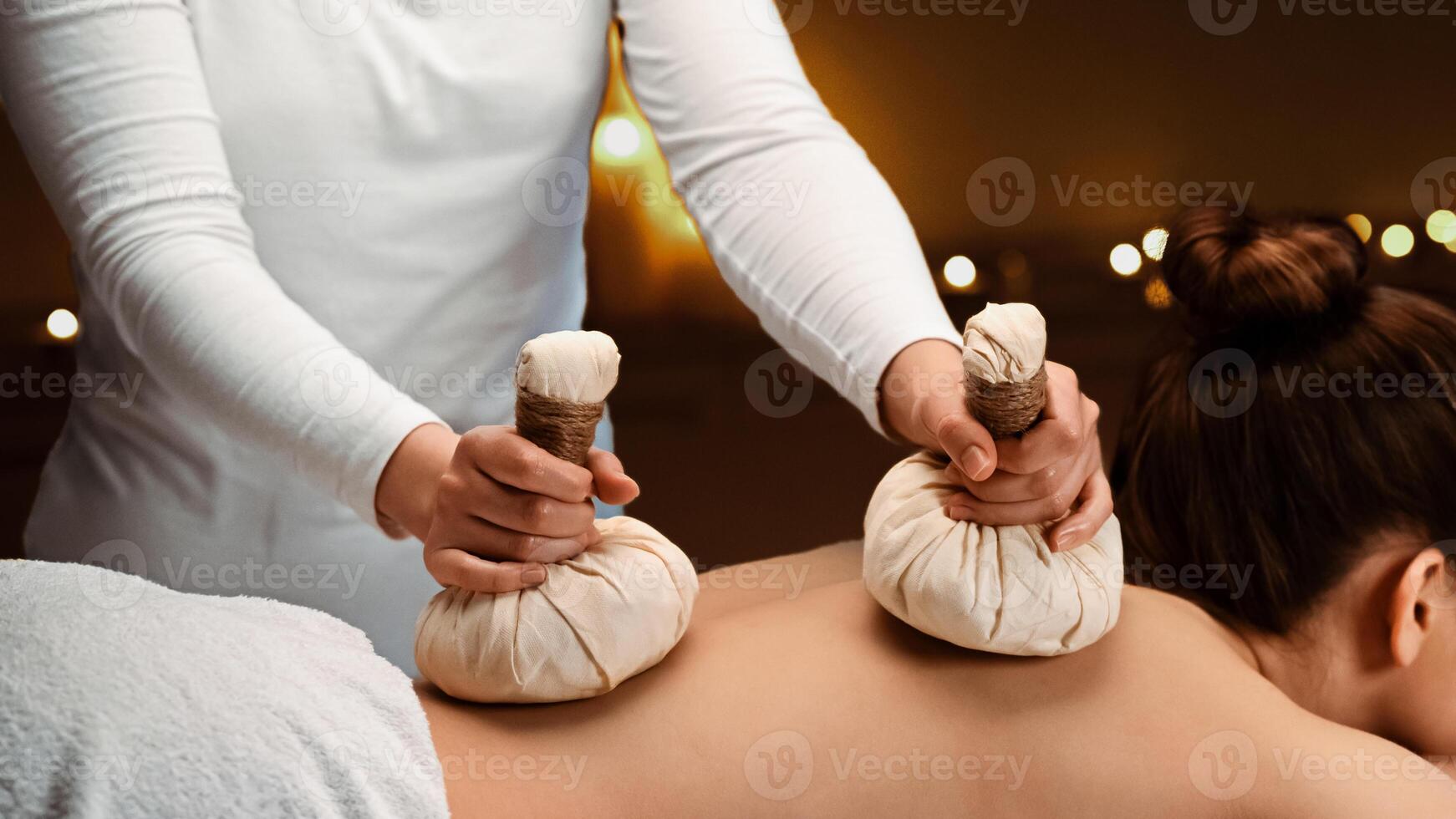 Herbal massage concept. Unrecognizable spa attendant making body massage for young lady with herbal compress balls, luxury spa interior. Woman enjoying herbal massage and aromatherapy at salon photo