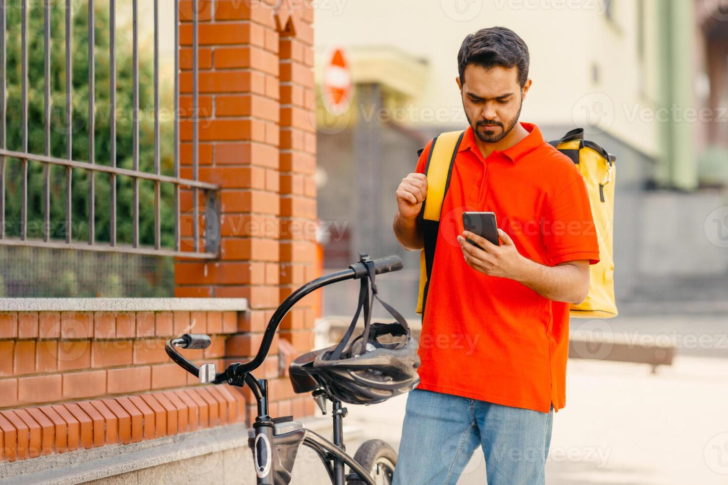 GPS map, coordinates of client's address, delivery service and online order. Concentrated young arabian male courier in orange t-shirt with bicycle and big thermo bag looks in phone, empty space photo