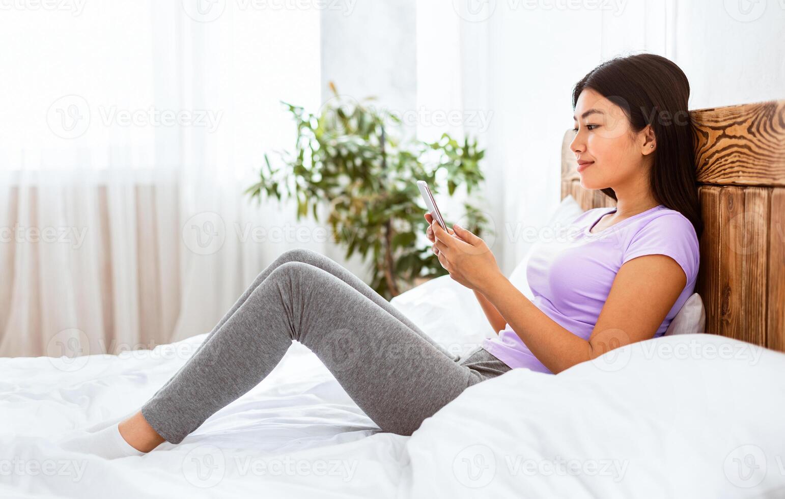 Asian Woman Using Phone Sitting In Bed At Home Relaxing On Weekend. Mobile Application Concept photo