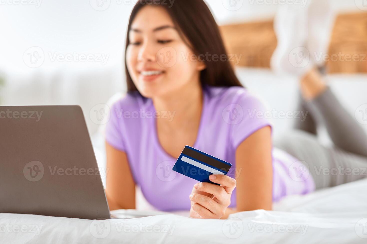 Asian Woman Doing Online Shopping Using Laptop And Credit Card Lying On Bed At Home On Weekend. Selective Focus photo