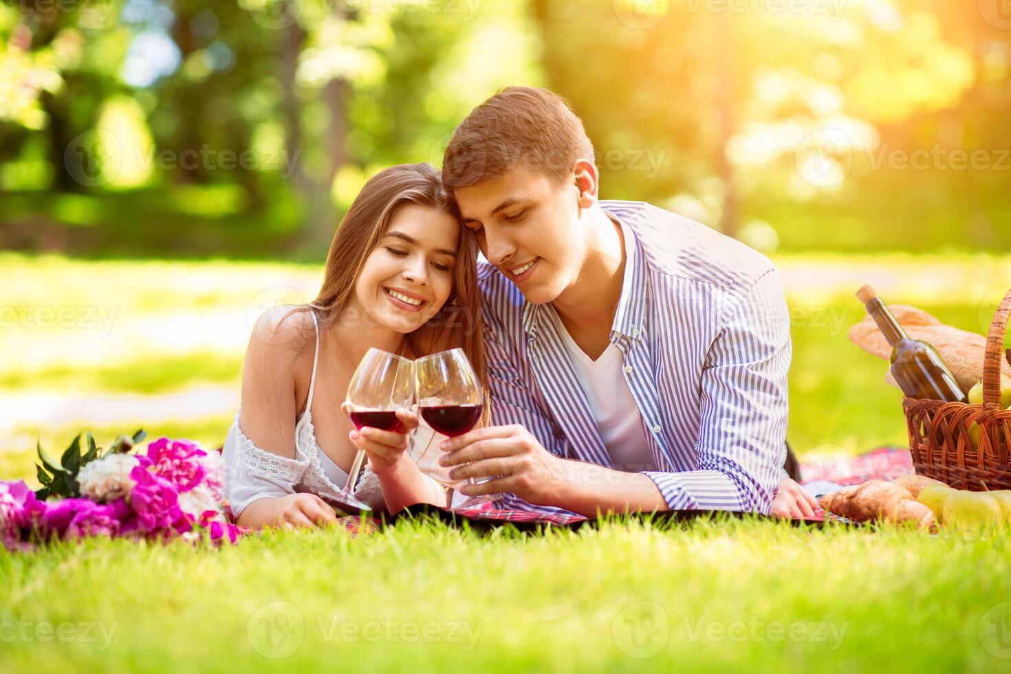 Affectionate young couple lying on picnic blanket with glasses of red wine, outdoors photo