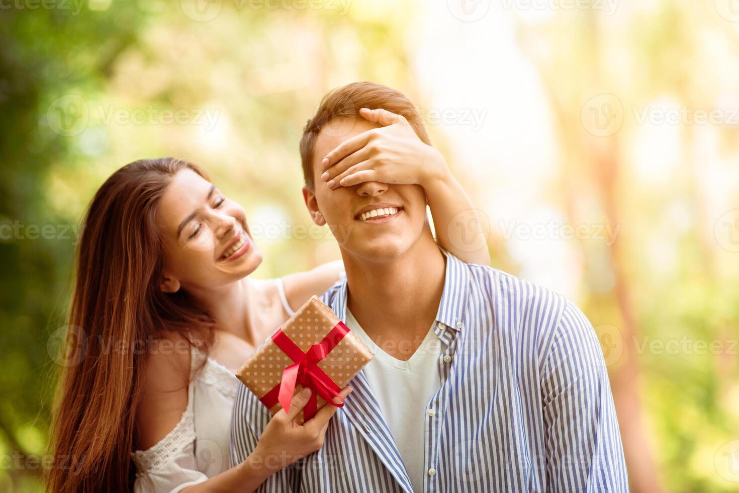 Attractive young girl closing her boyfriend's eyes and surprising him with birthday gift at park photo