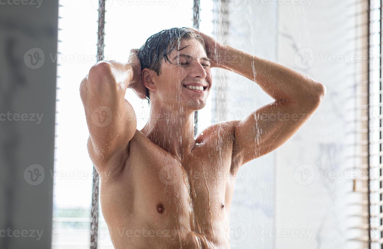 Selfcare And Wellness Concept. Portrait Of Smiling Handsome Young Man Taking A Shower Standing Under Falling Water Drops Washing Naked Body And Head In Modern Bathroom At Home. Male Model photo
