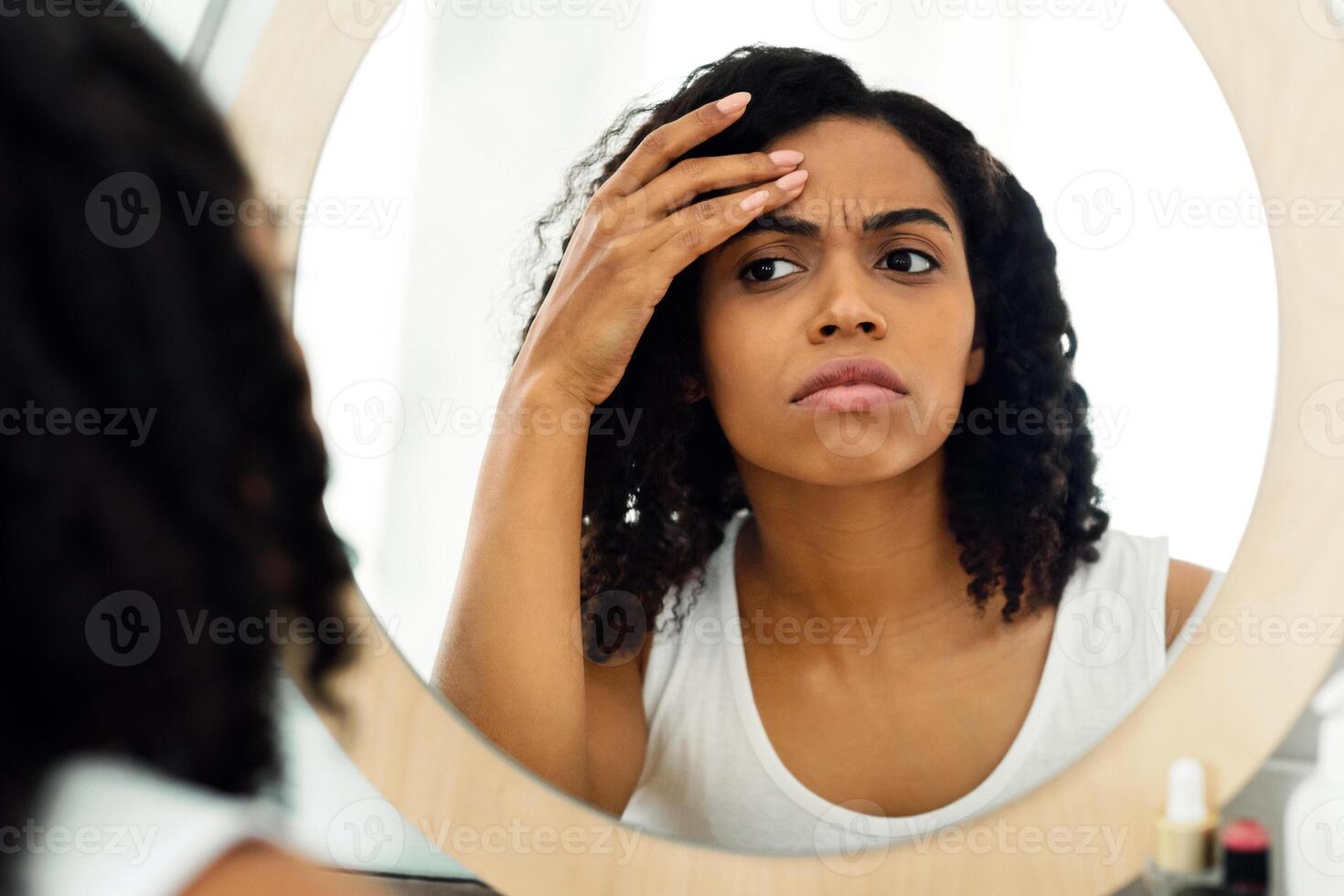 Fine lines and wrinkles. Concerned black woman looking in mirror checking aging signs on her face, selective focus photo
