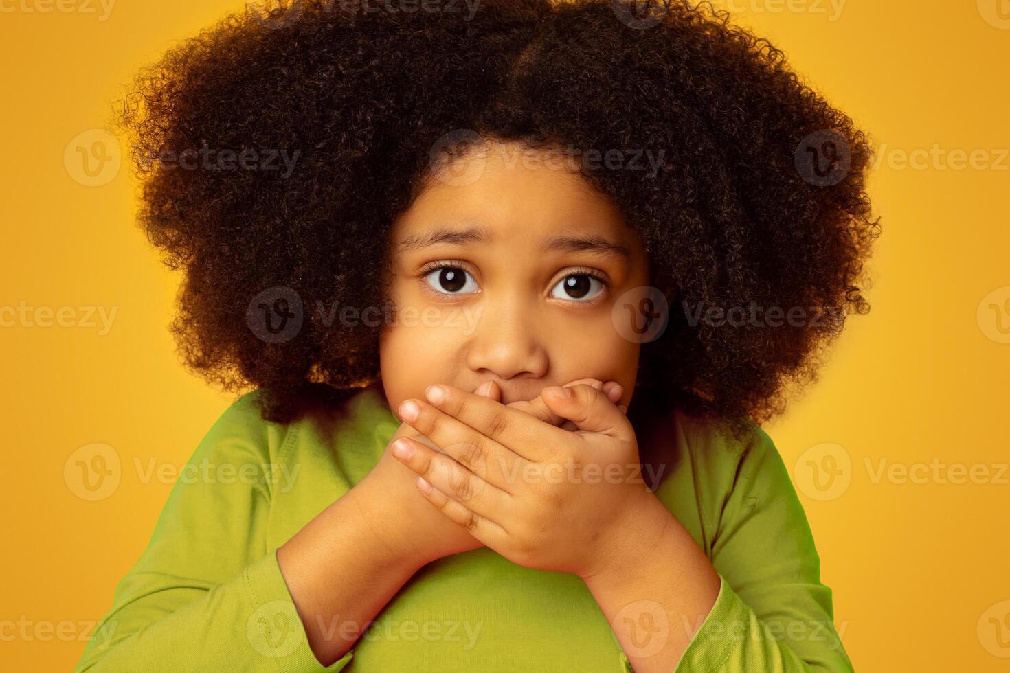 Keep Silent. Frightened little girl covering mouth with hands and looking at camera with scared face expression, posing over yellow background photo