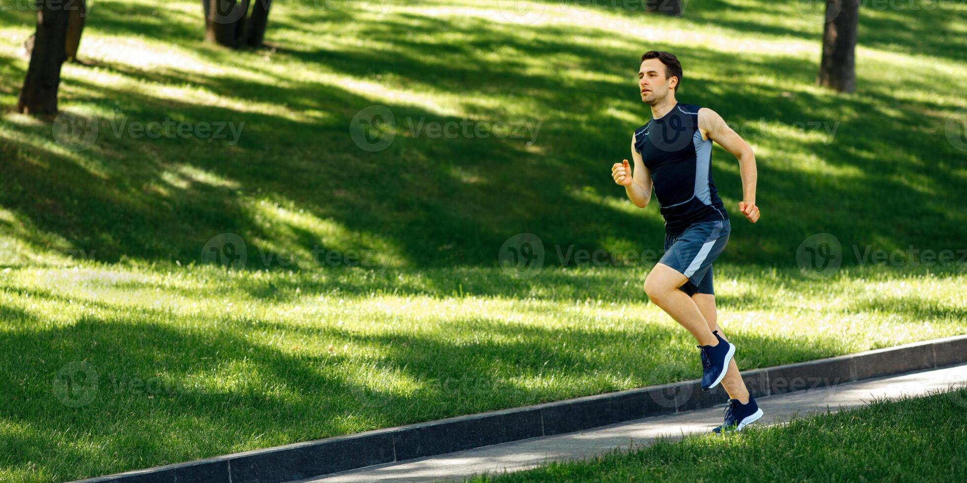 Urban sport and outdoor training. Muscular guy in sportswear in sneakers with fitness tracker runs at path in park, free space photo