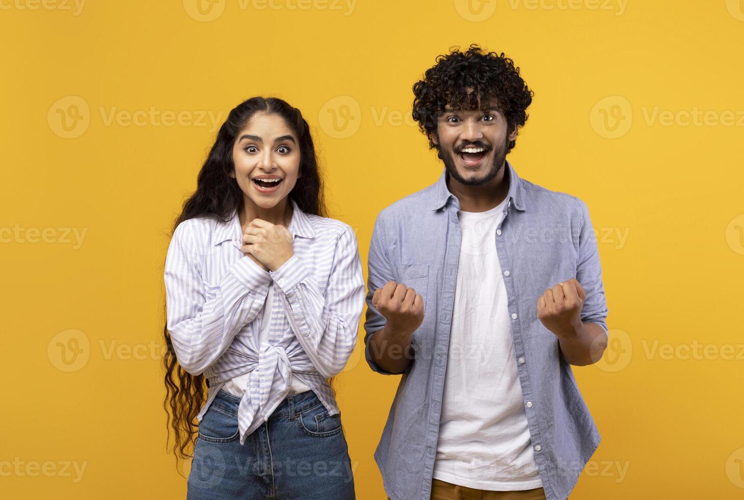 Wow, yes. Portrait of overjoyed indian man and woman cheering and shaking clenched fists, looking at camera in excitement. Happy couple celebrating win over yellow studio background wall photo