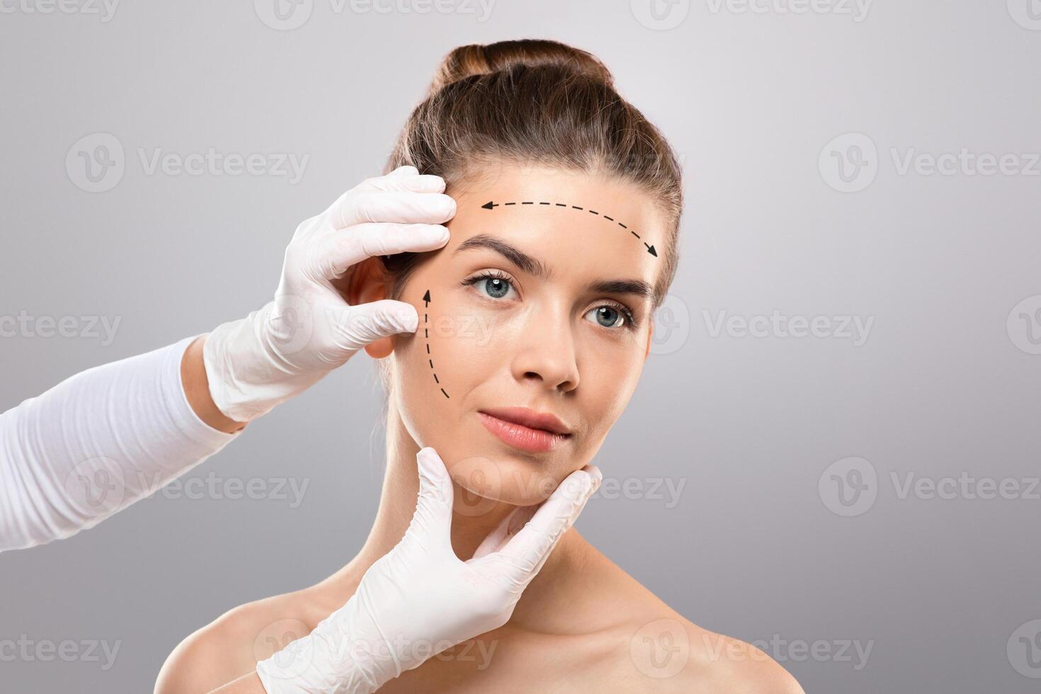 Concept of plastic surgery. Young lady getting treatment at cosmetology clinic, changing her appearance. Plastic surgeon making marks on beautiful woman face, grey studio background, copy space photo