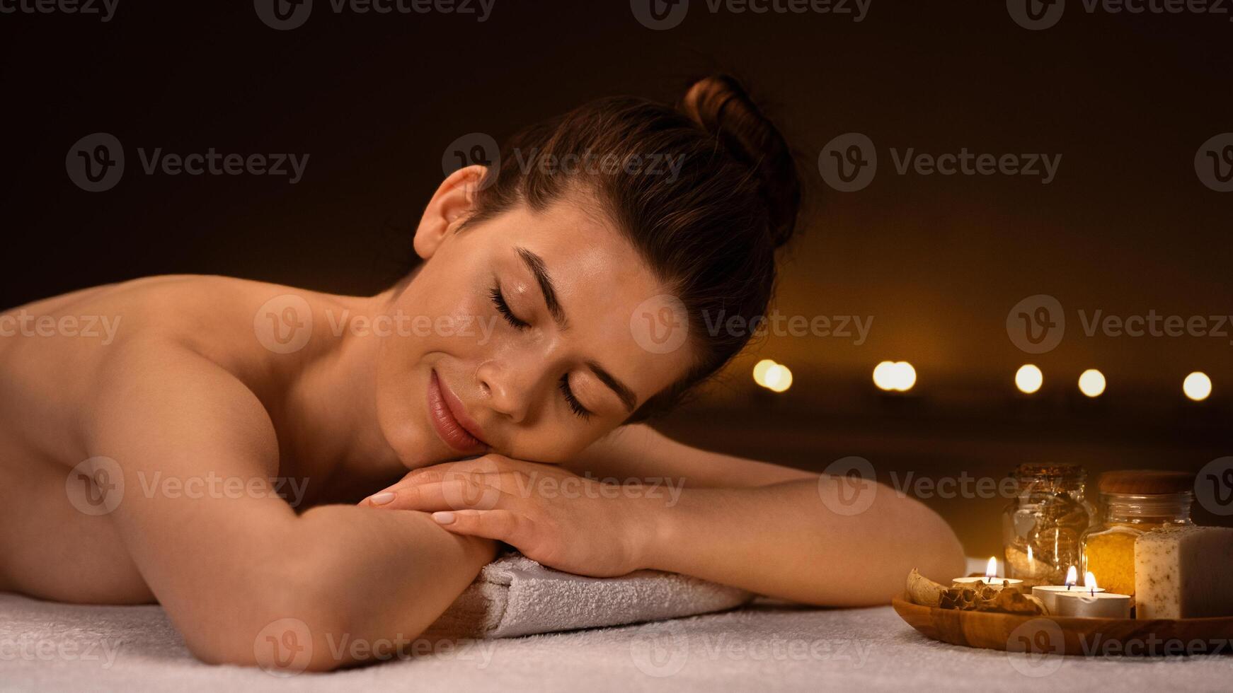 Side view of tranquil young woman spending day at spa salon decorated with candles, getting aroma session with relaxing full body massage, panorama with copy space. Spa, massage, relaxation concept photo