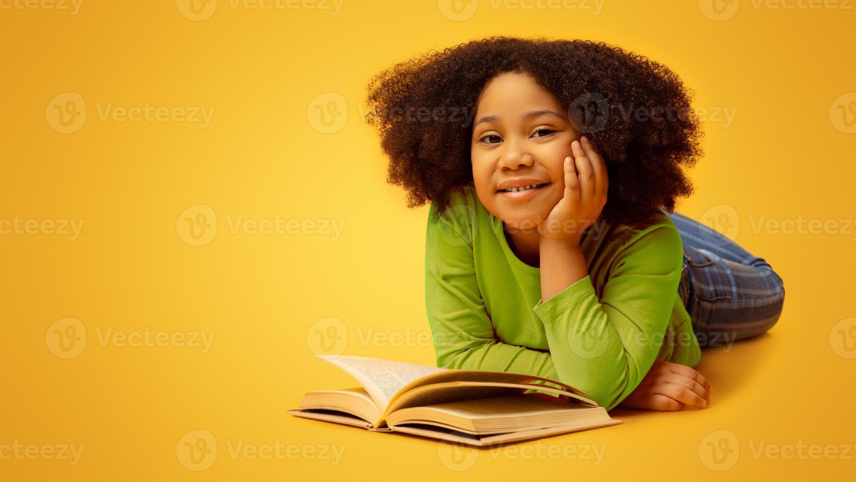 Cute Little African American Girl Reading Book On Floor In Studio And Smiling At Camera, Lying On Yellow Background, Free Space photo