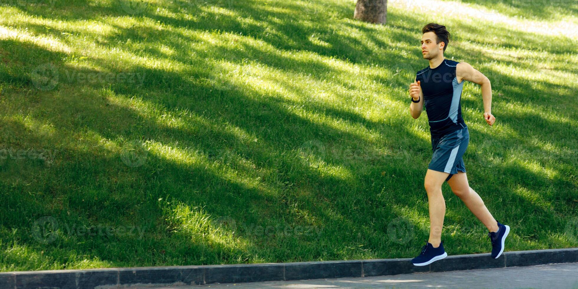 Muscle workout and jogging. Young man in sportswear with fitness tracker runs along track at green park, free space photo