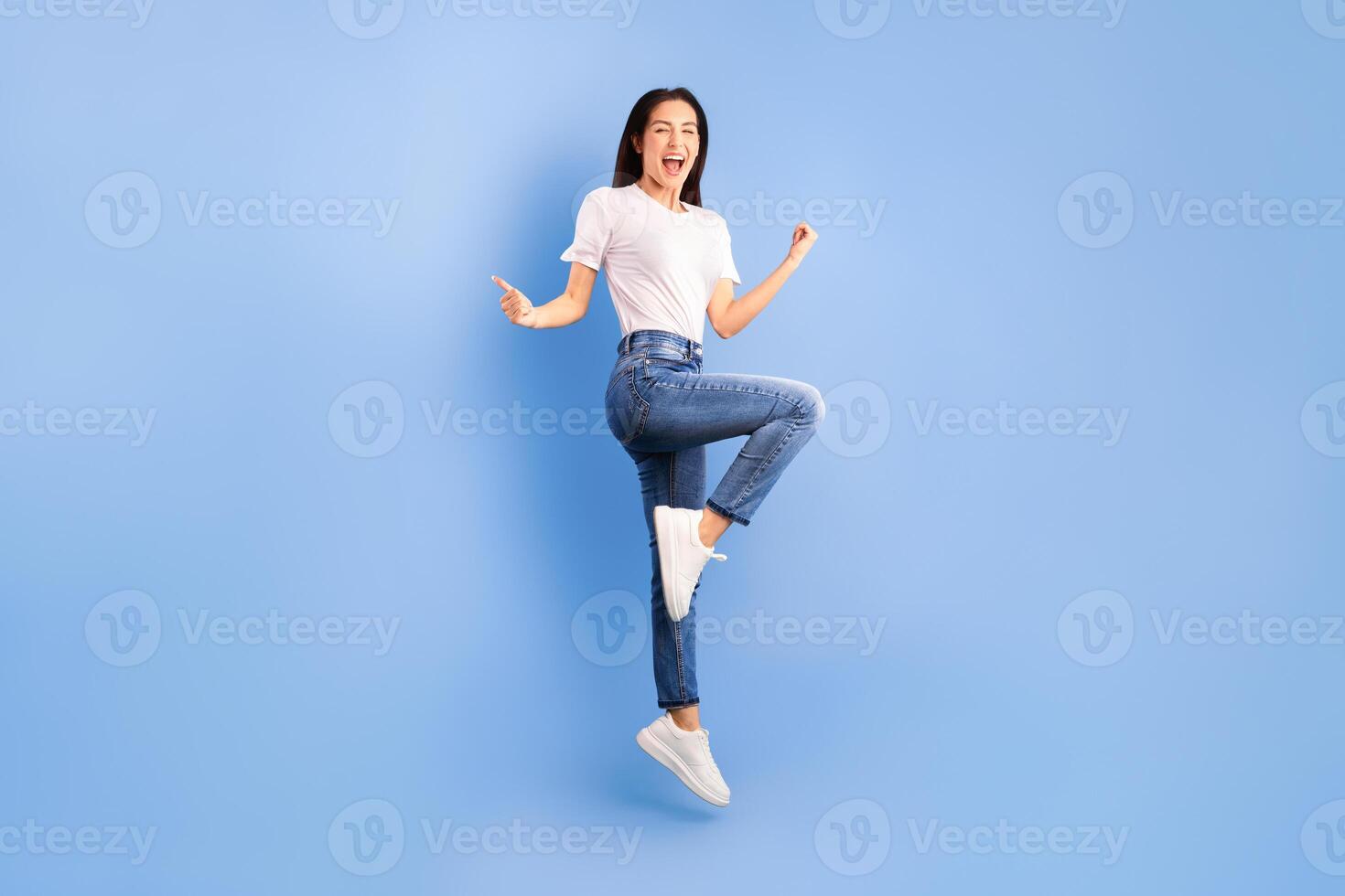Yeah. Excited lady jumping and showing thumbs up gesture isolated on blue studio wall, full length photo