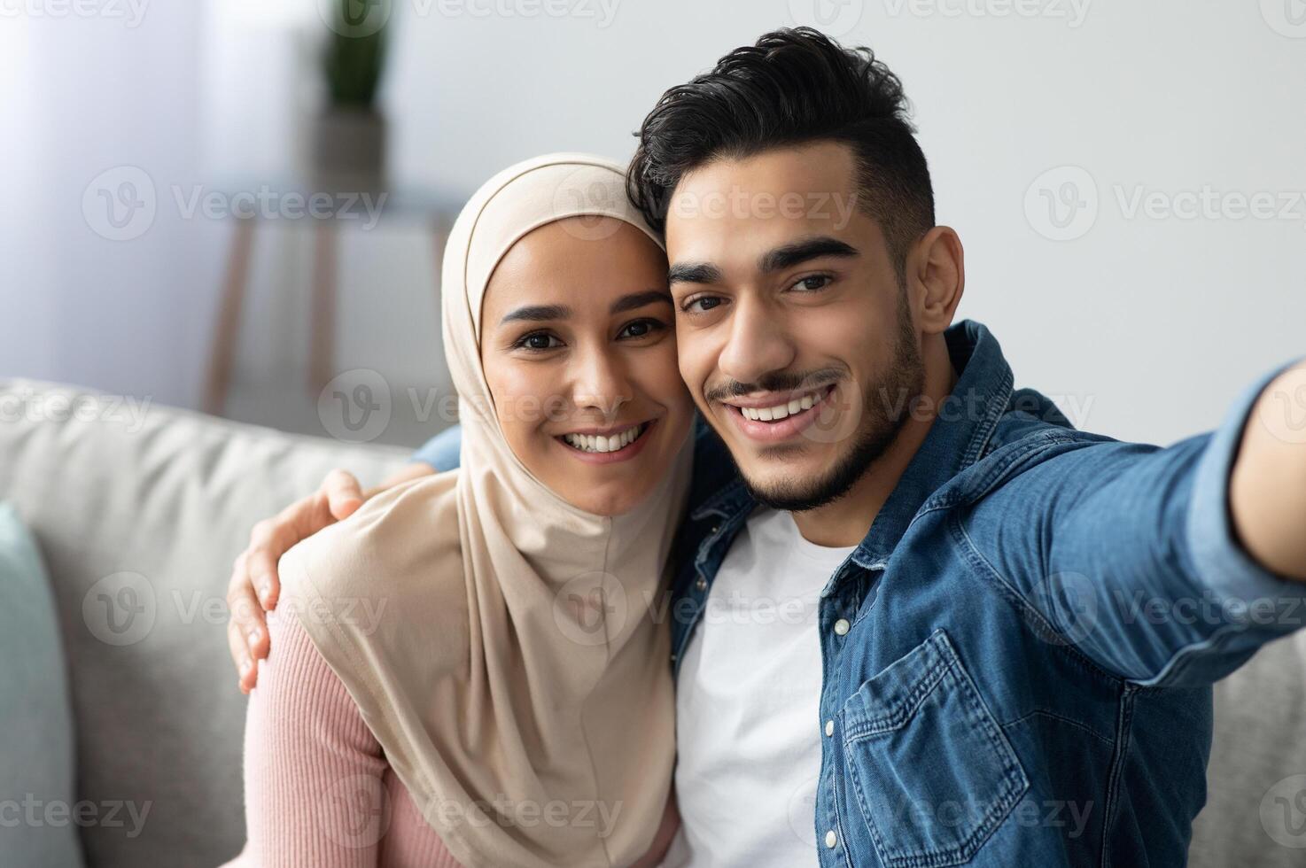 Beautiful muslim man and woman in hijab taking selfie from home, copy space. Smiling middle eastern young couple hugging while having call, sitting on sofa in living room photo