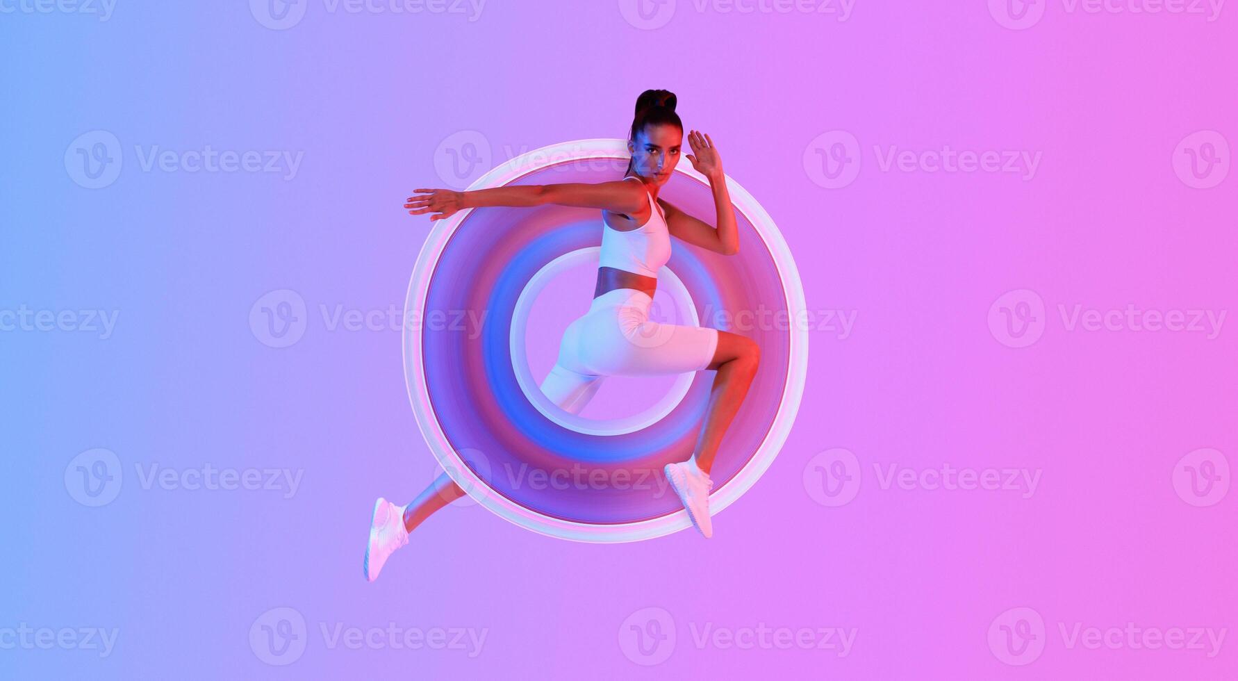 Athletic Woman Jumping Posing In Mid-Air Looking At Camera Exercising Over Pink And Blue Neon Background In Studio. Full Length, Panorama With Empty Space photo