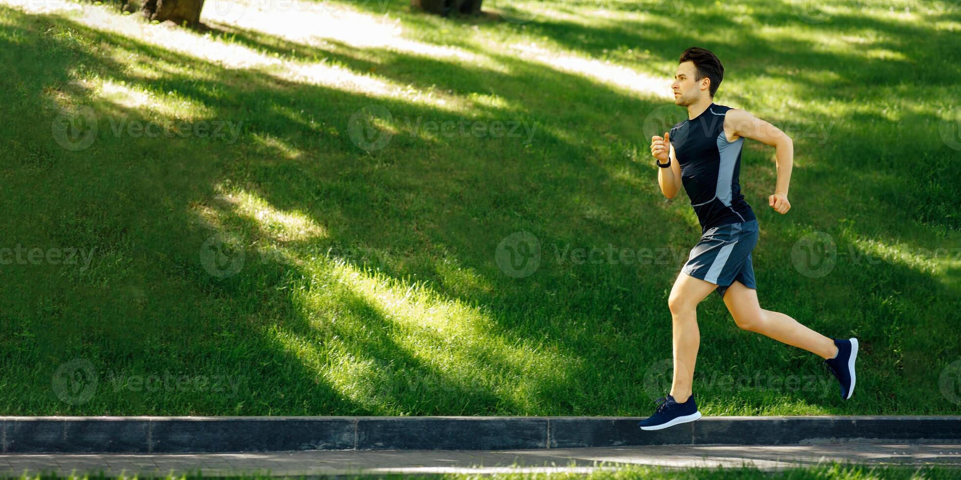 Sportsman in park. Guy in sportswear with sneakers and fitness tracker runs at track, side view, free space photo