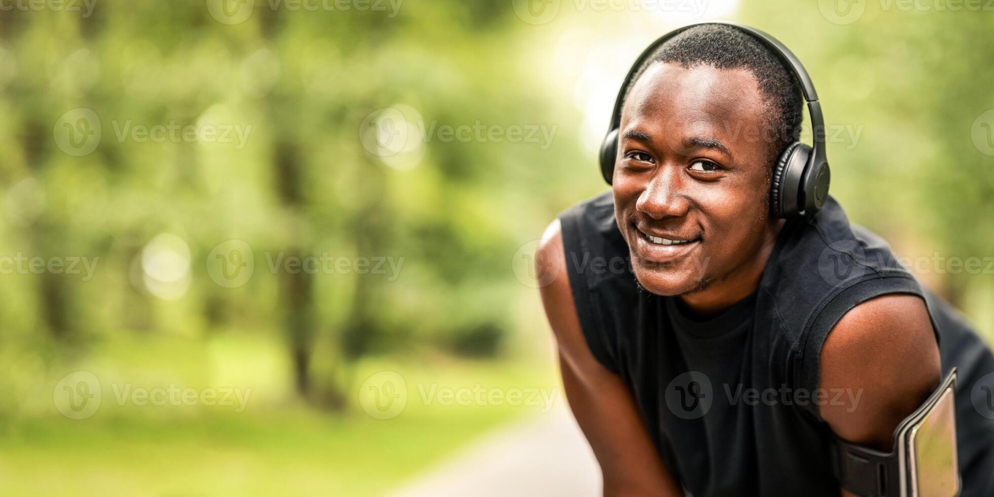 Closeup of cheerful black jogger with wireless headset resting during training at park, empty space photo