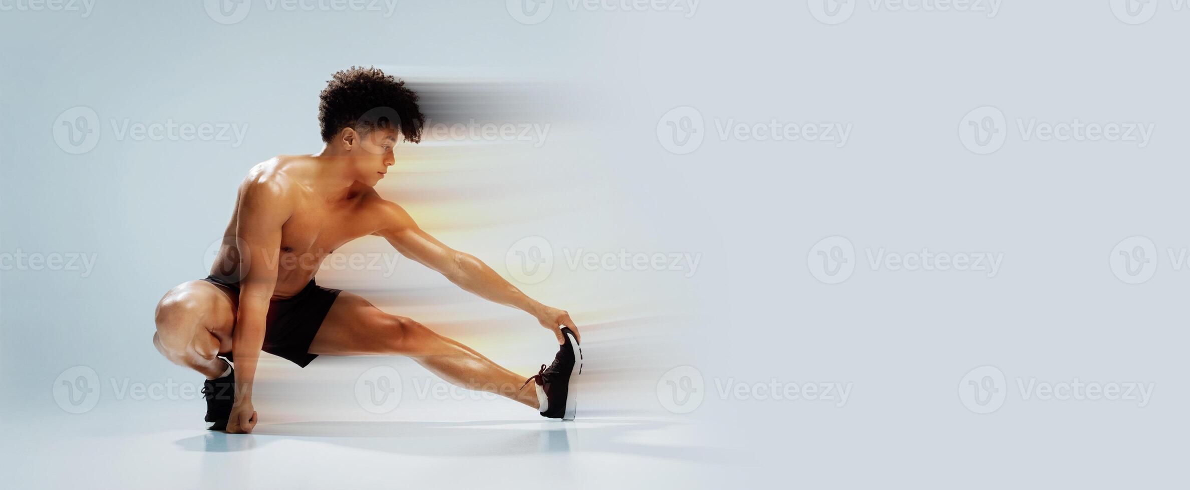 Fitness workout. Young shirtless athletic man exercising in extended leg squat pose, stretching legs and touching toes, demonstrating flexibility and strength over gray studio background photo