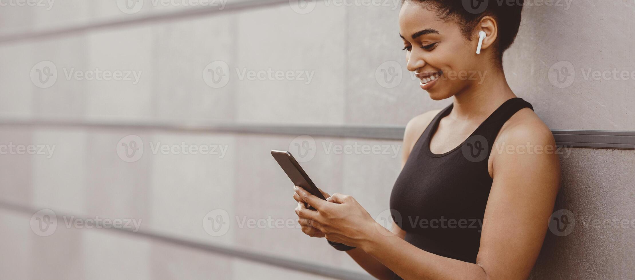 Start training and music for jogging. Smiling african american girl with wireless headphones looks in smartphone on gray wall background, close up, panorama, free space photo