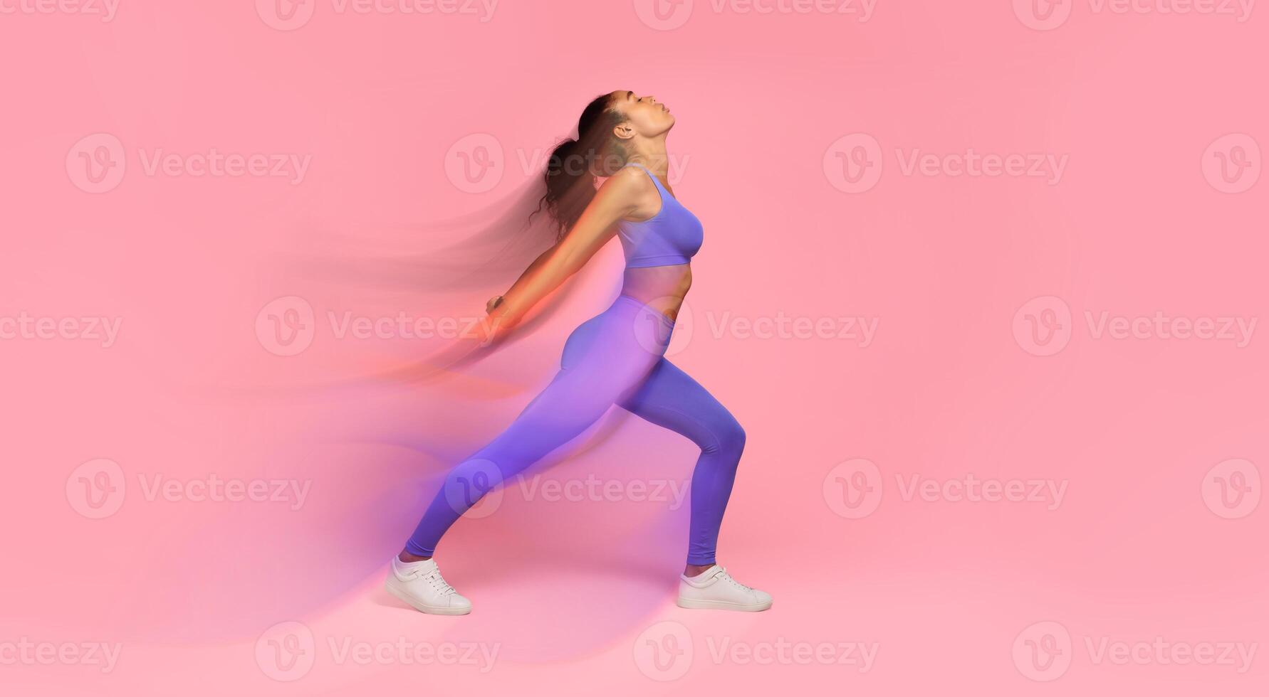Young African American woman in fitwear performing backbend in lunge position, stretching back muscles in bend, over pink studio backdrop. Fitness routine and workout in gym. Full length photo