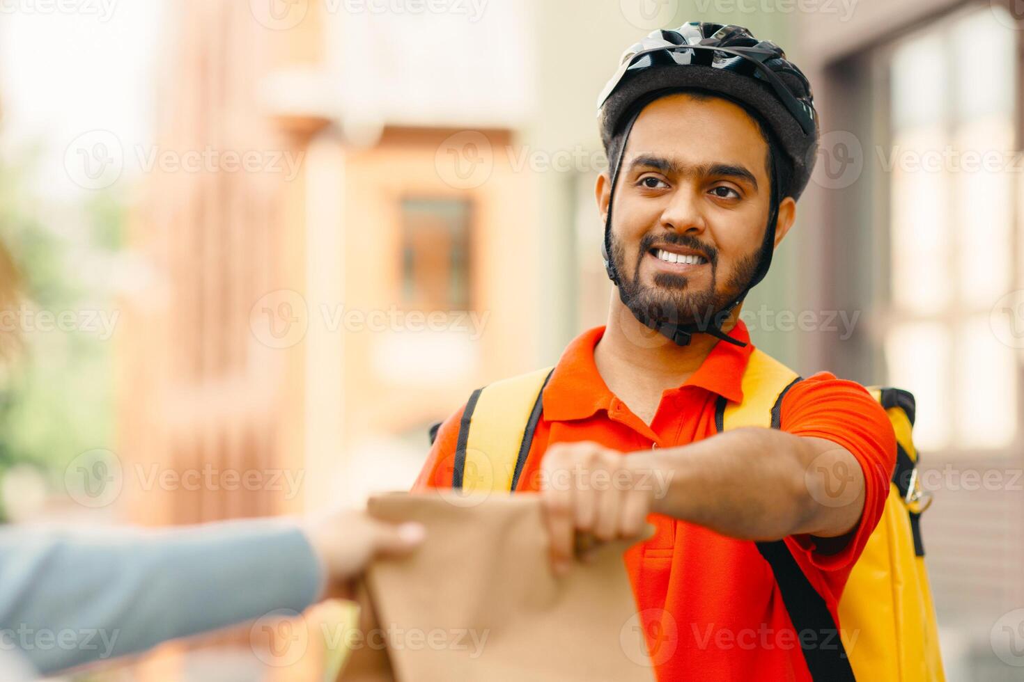 Takeaway restaurant food delivery. Portrait of young courier with beard in protective helmet and backpack, gives paper bag to girl, close up, free space photo