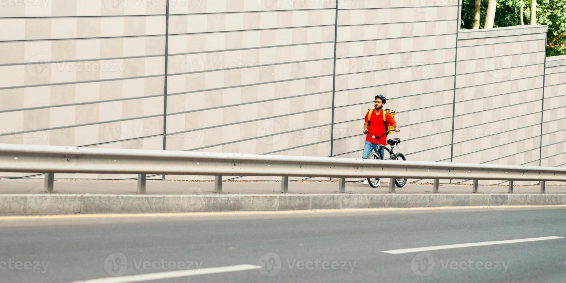 Delivery route. Courier in helmet, with yellow backpack and bicycle walks along the track on street to client on gray wall backgroundin town, free space photo