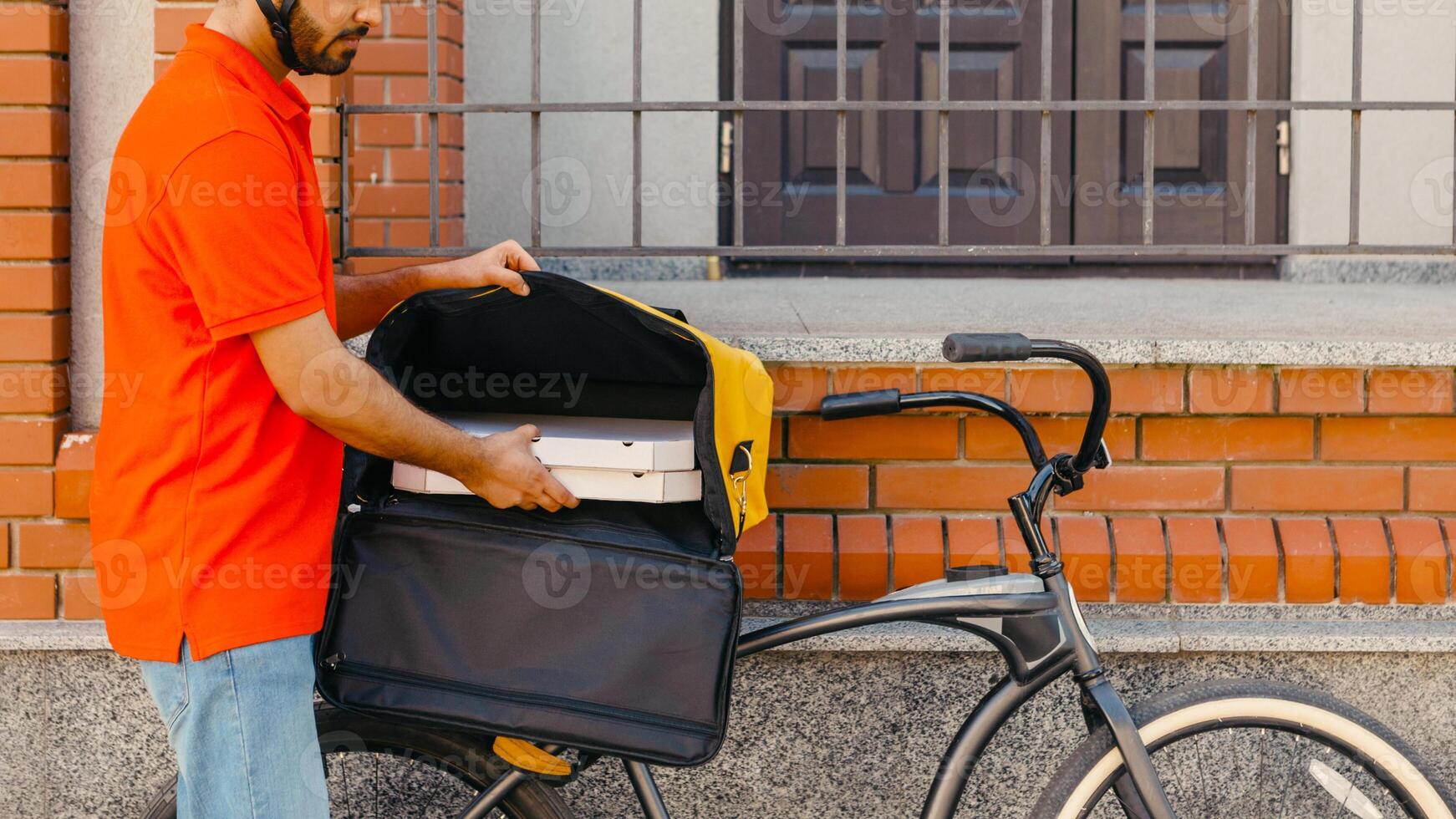 Delivery home and office. Young delivery man in helmet takes out pizza from big yellow backpack on bicycle, near front door, side view, free space photo