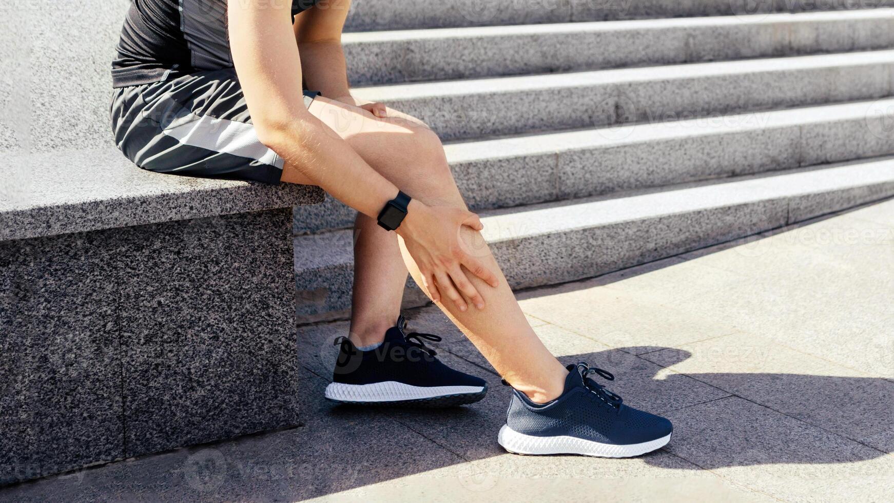 Leg injury while jogging. Guy with expression of pain on face, sits and rubs leg on street, free space photo