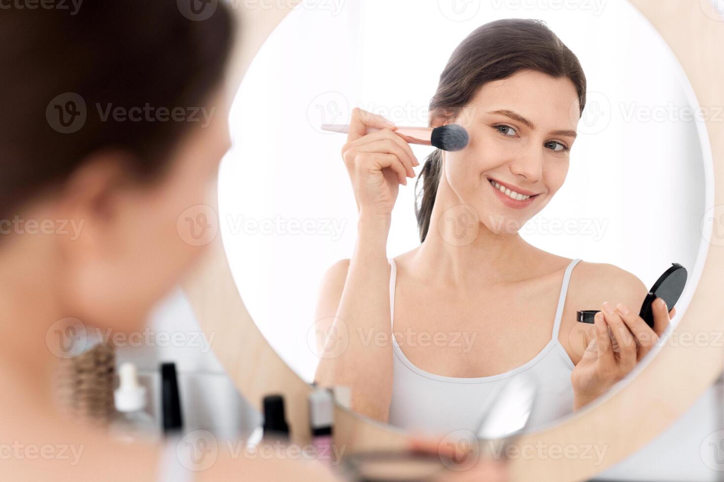 Happy pretty young woman applying blush on her face, sitting in front of mirror, home interior, copy space. Smiling attractive young lady doing makeup before going out, shot over shoulder in bedroom photo